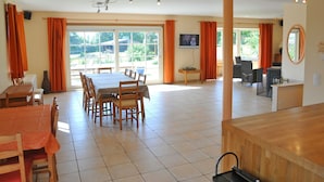 A spacious dining area with a large table and chairs, a television mounted on the wall, and a view of the outdoors through large windows.