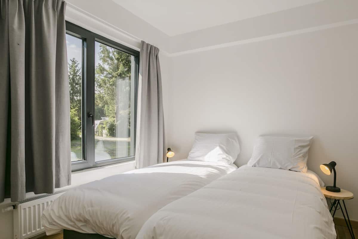 Une chambre avec une grande fenêtre, un lit recouvert de linge de lit blanc, deux lampes de chevet et une petite table ronde.