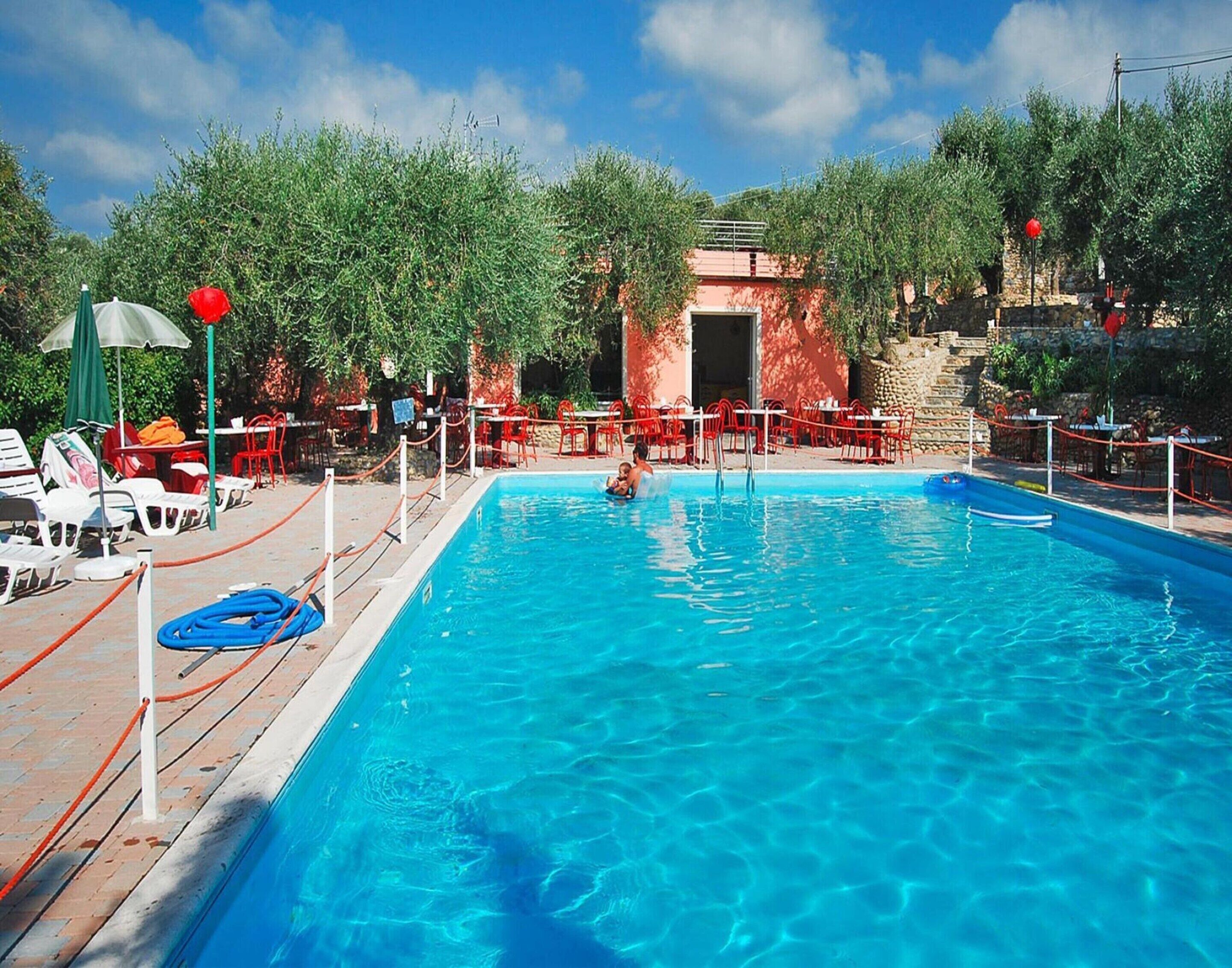 Una piscina con un bambino che nuota, circondata da sedie e ombrelloni rossi.
