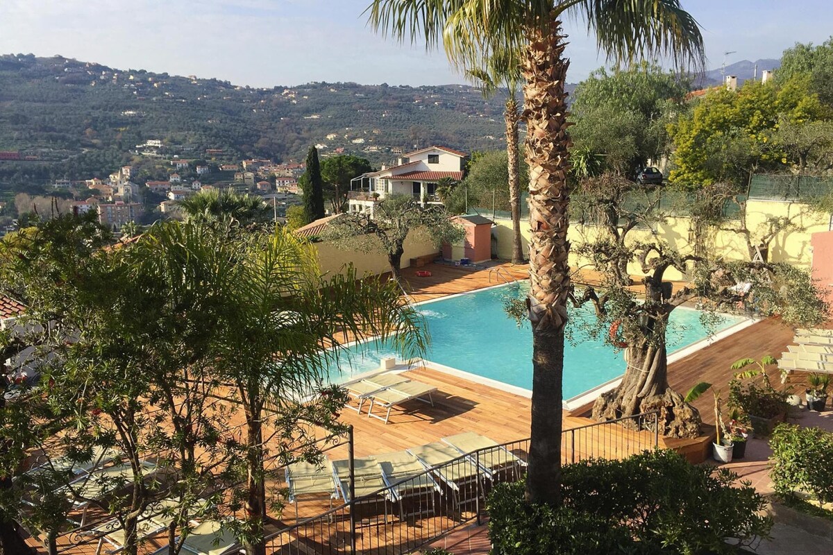 Un espace piscine avec des chaises longues, un palmier et une vue sur une ville située sur une colline.