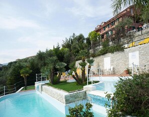 Μια πισίνα με πέτρινο τοίχο και πλούσια βλάστηση, ένα πέτρινο κτίριο με μπαλκόνια και ένας καθαρός ουρανός.