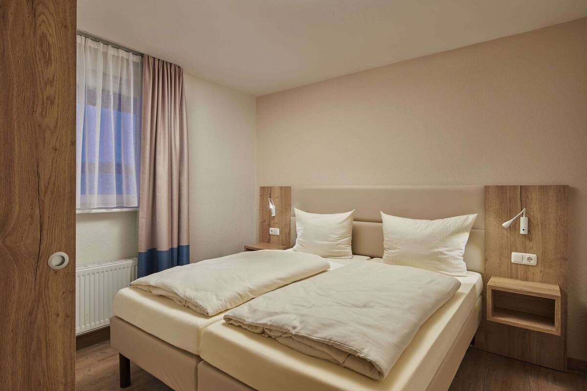 A double bed with two pillows, a bedside table, a wall-mounted lamp, and a window with curtains.