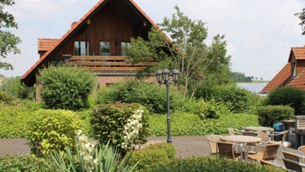 Eine Holzhütte mit Balkon, umgeben von üppigem Grün und einer Terrasse mit Sitzgelegenheiten.