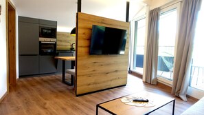 Un espacio habitable moderno con un separador de madera, un televisor de pantalla plana, una zona de cocina y un balcón con cortinas.