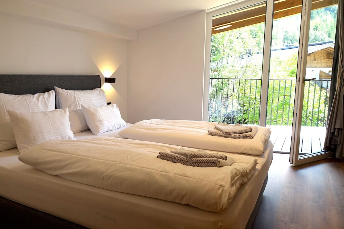 Ein Schlafzimmer mit zwei Betten, einem dunklen Kopfteil und Blick auf Grün durch eine Schiebetür.