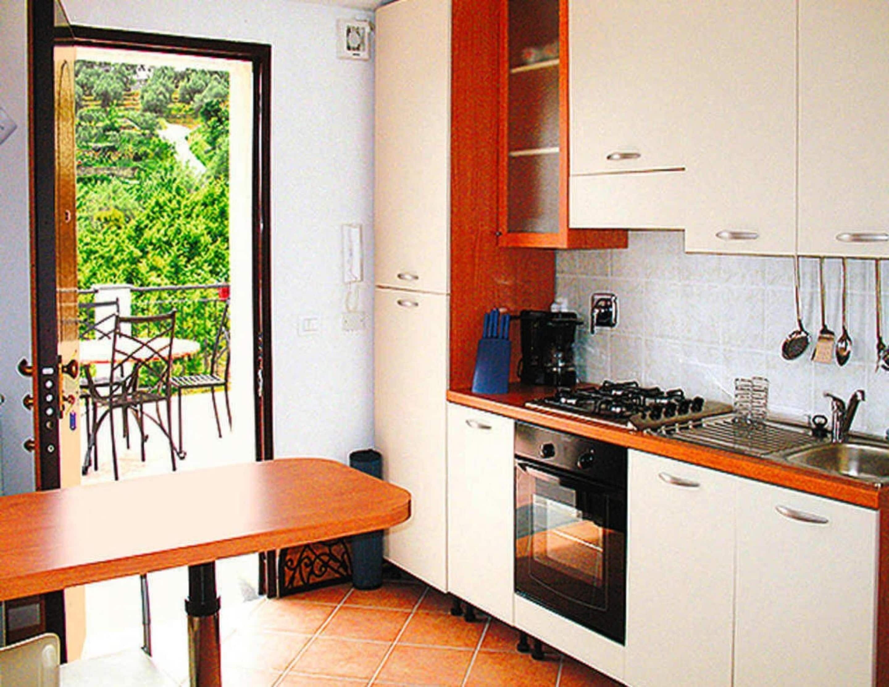 A compact kitchen with white cabinets, a stove, and a sink. There is a dining table with chairs outside the door.