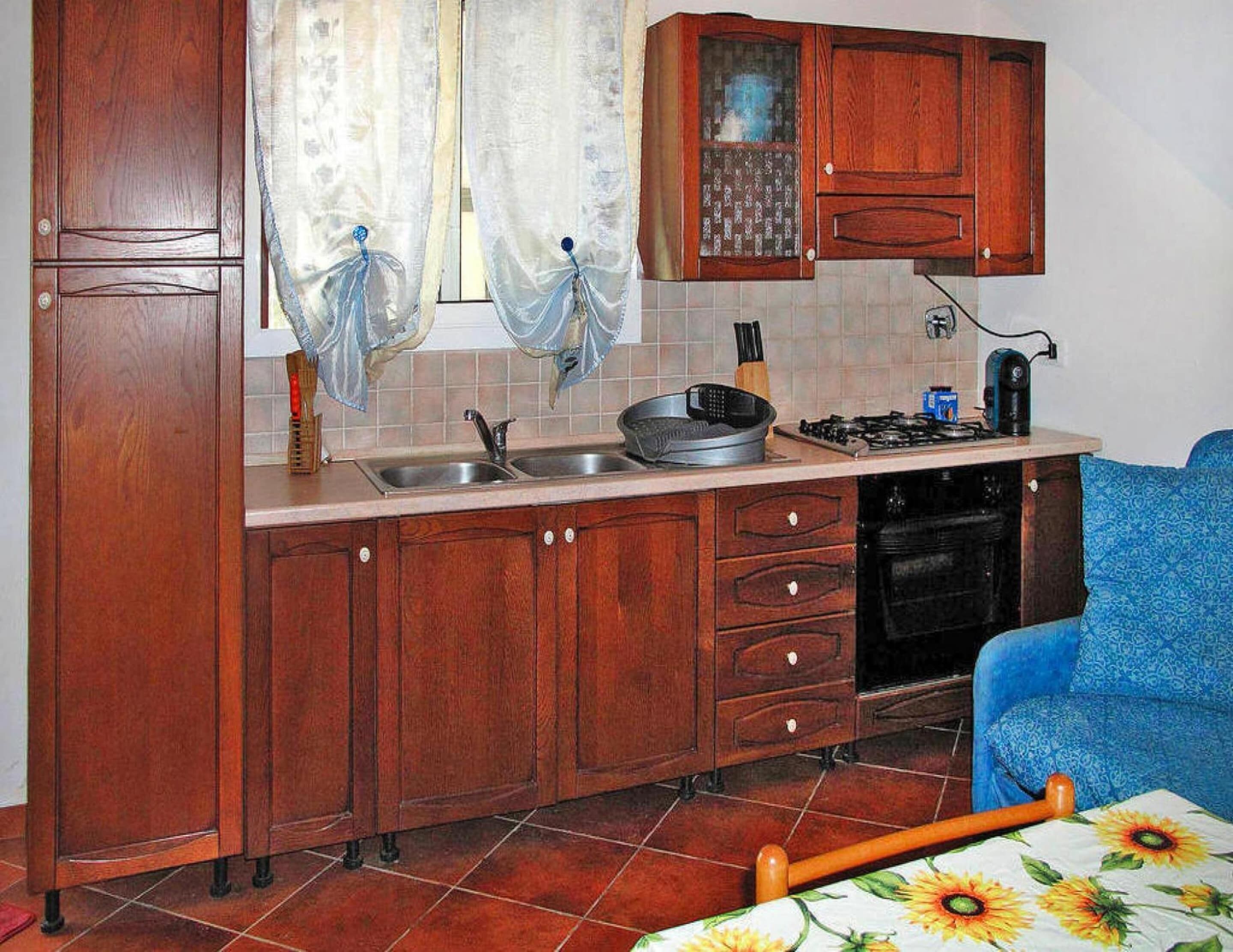 A compact kitchen with wooden cabinets, a sink, a stove, and a blue chair.