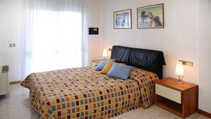 A bedroom with a checkered bedspread, two pillows, a wooden nightstand with a lamp, and two framed pictures on the wall. - Apartment in the Residence Delle Terme in Bibione Spiaggia (San Michele al Tagliamento)