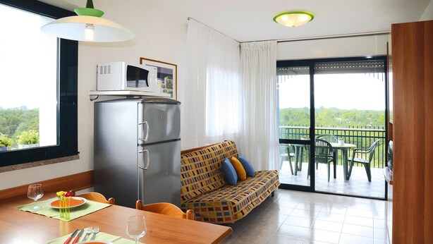 A dining area with a table set for two, a sofa, a refrigerator, and a balcony with outdoor furniture. - Apartment in the Residence Delle Terme in Bibione Spiaggia (San Michele al Tagliamento)