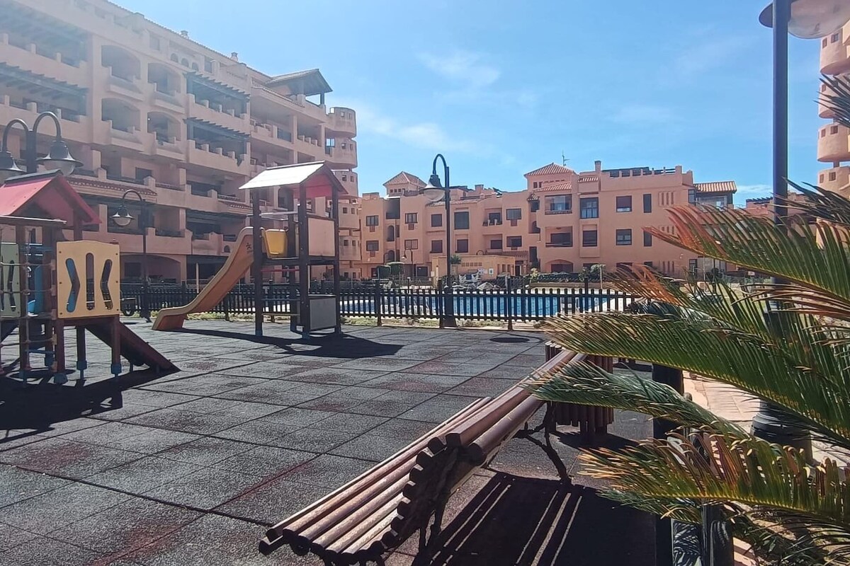 Une aire de jeux avec un toboggan et des structures d’escalade, entourée d’une zone délimitée par une clôture, où se trouvent des bancs et des palmiers.