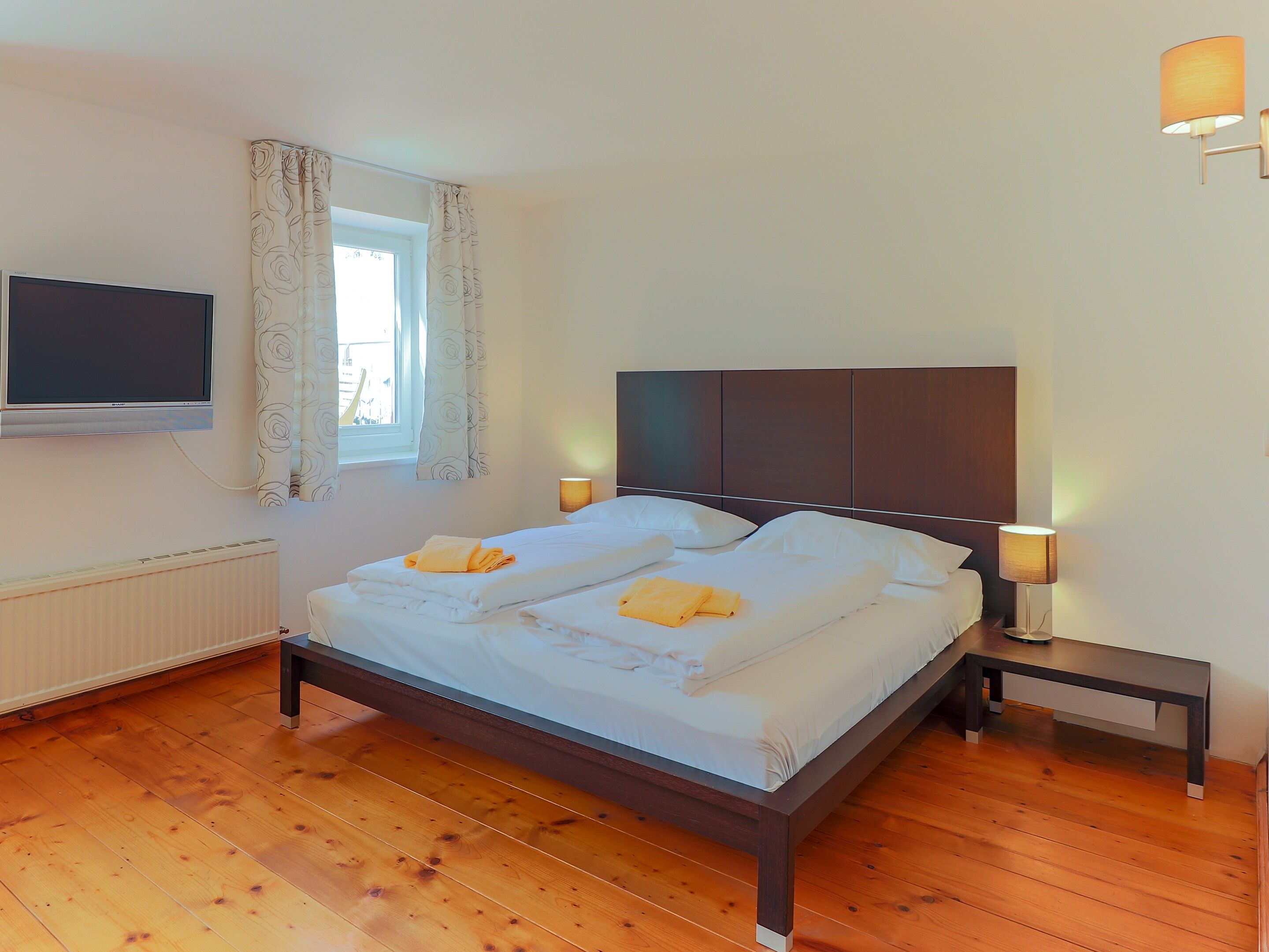 A bedroom with a bed, two bedside tables, a TV mounted on the wall, and a window with curtains.