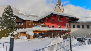 A red building with a steeply pitched roof, surrounded by snow-covered trees and mountains.