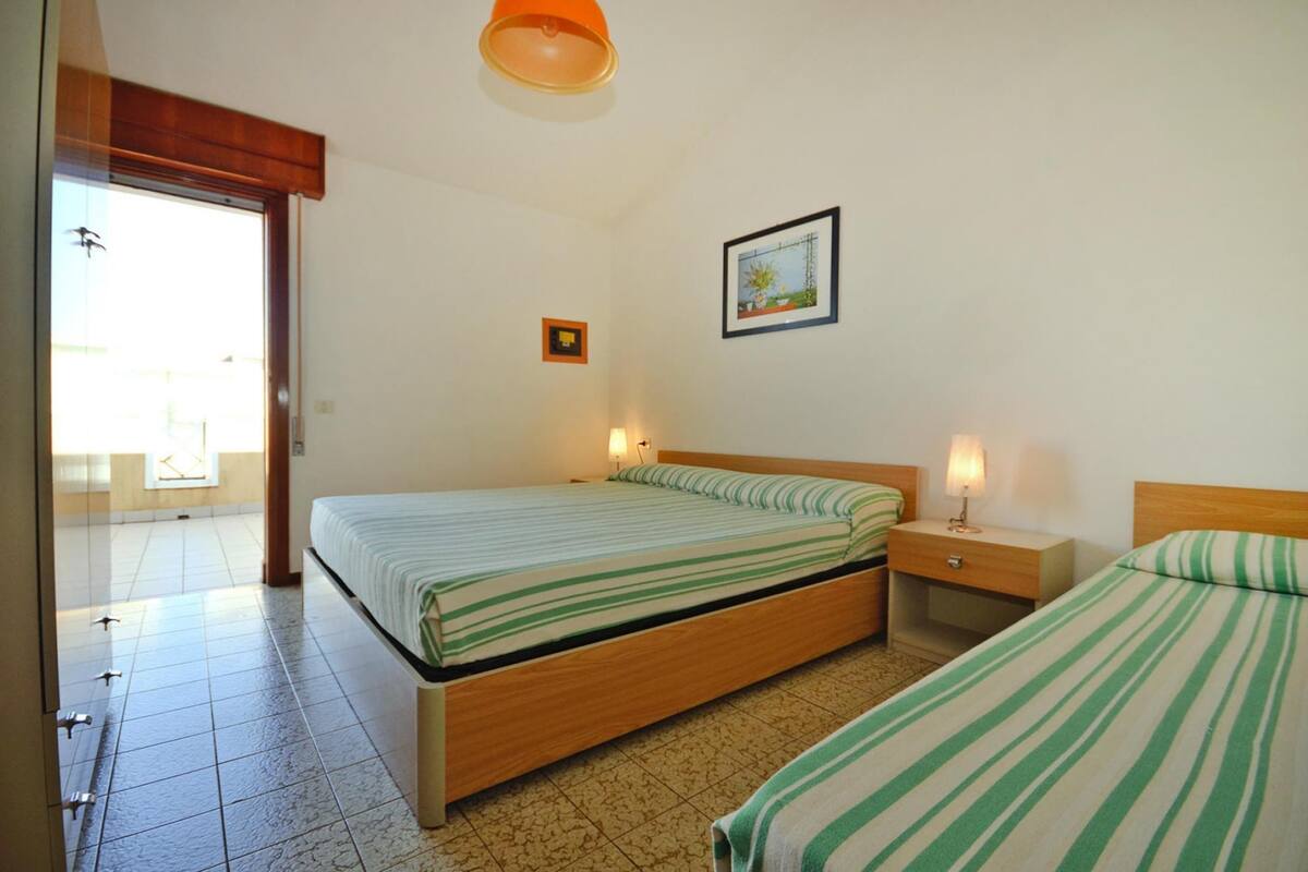 A bedroom with two beds, a wooden headboard, a nightstand with a lamp, a framed picture on the wall, and a sliding glass door leading to a balcony.