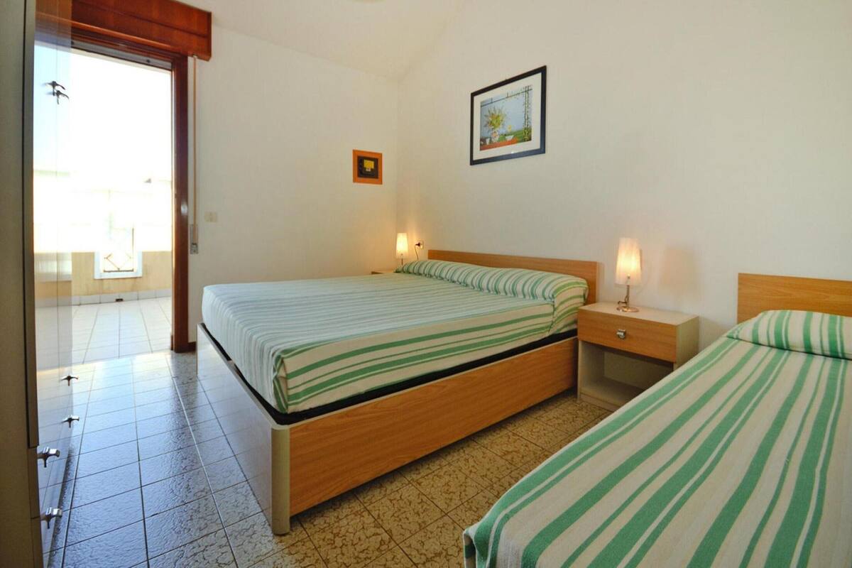 A bedroom with two beds, a wooden headboard, a bedside table with a lamp, a framed picture on the wall, and a door leading to a balcony.