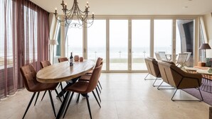 A spacious dining area with a large wooden table and modern chairs, a chandelier, and a view of the ocean through large windows. - Luxury Apartment in a Residence Right on the Boulevard and the Beach (Egmond aan Zee)