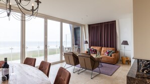 A spacious living area with a balcony overlooking the ocean, a dining table with chairs, a sofa with cushions, and a fireplace. - Luxury Apartment in a Residence Right on the Boulevard and the Beach (Egmond aan Zee)