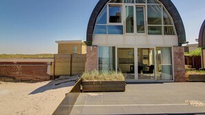 A modern two-story house with a curved roof, large glass windows, and a patio area with a wooden planter. - Luxury Apartment in a Residence Right on the Boulevard and the Beach (Egmond aan Zee)