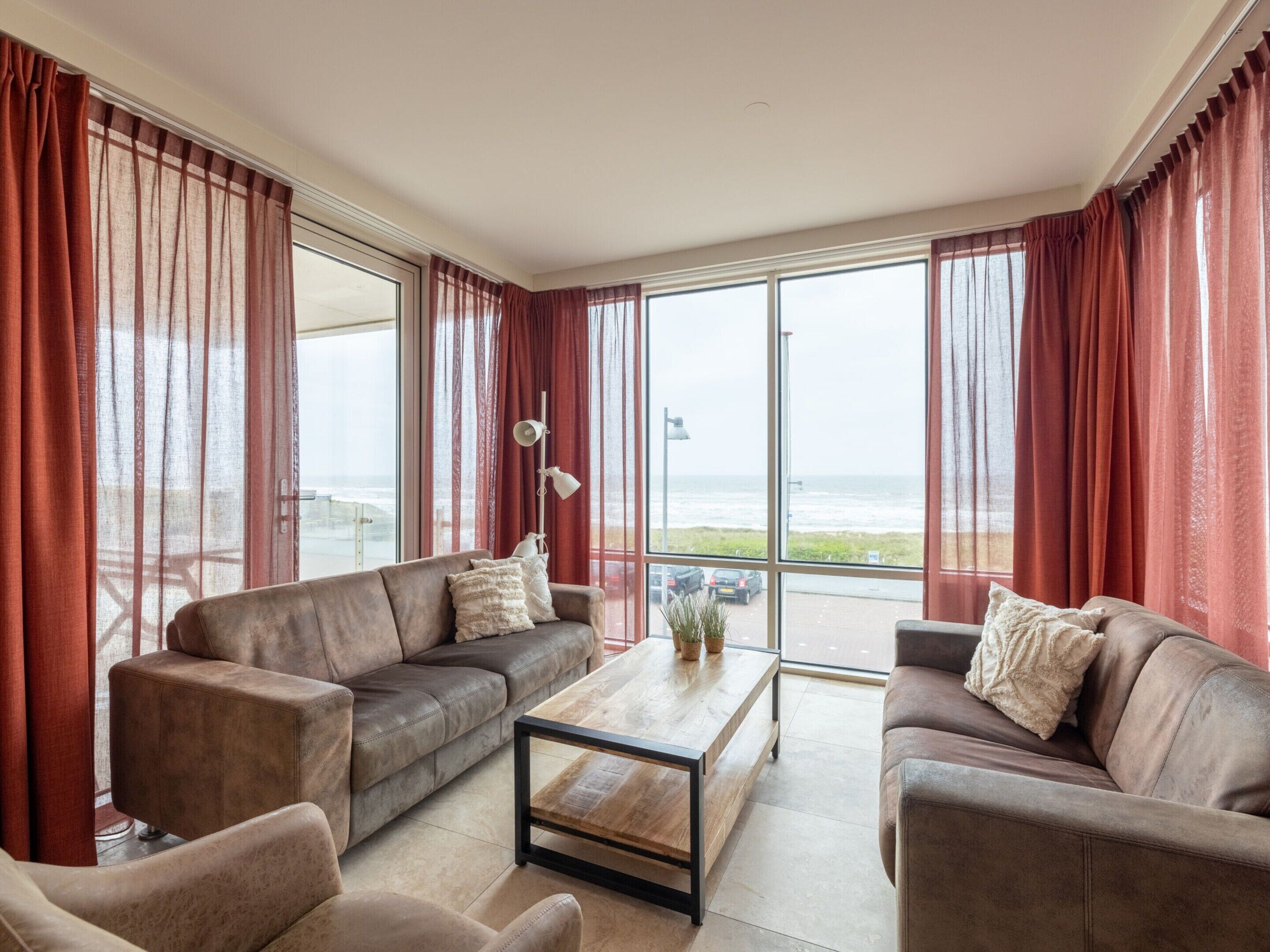 Un salón amplio con dos sofás y una mesa de centro, ventanas grandes con cortinas rojas y vista al mar.