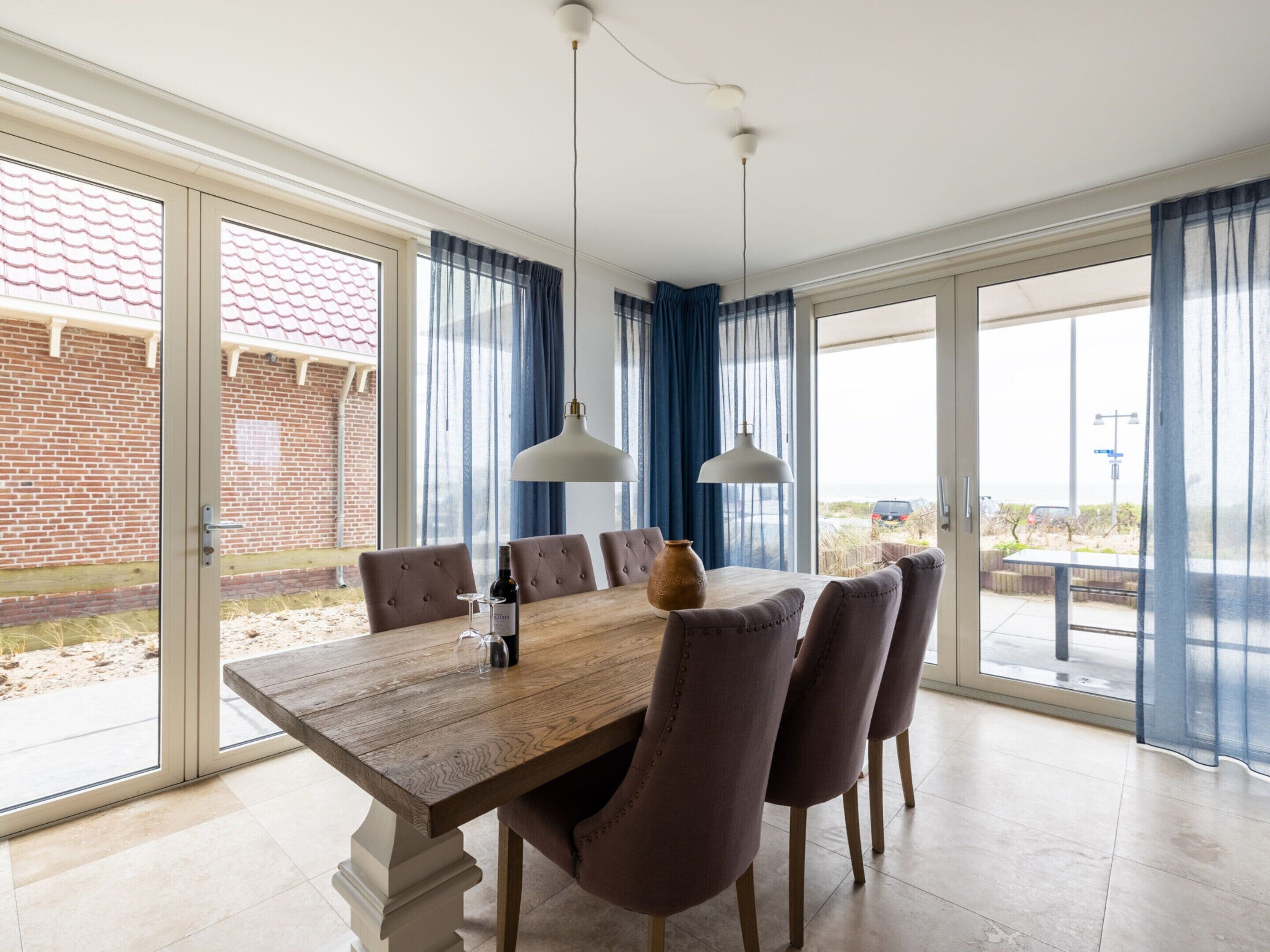 A dining area with a wooden table, six chairs, two pendant lights, and a bottle of wine.