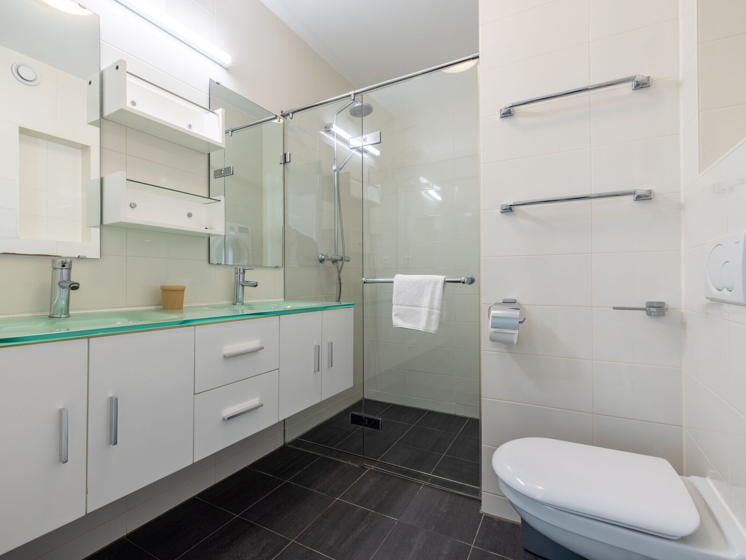 Une salle de bain moderne avec une douche à parois de verre, des toilettes blanches et un lavabo surmonté d’une plaque de verre.