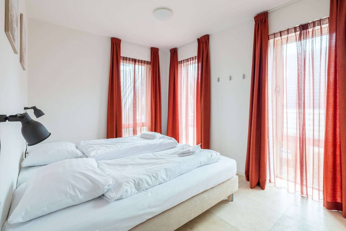 Una cama doble con sábanas blancas, una lámpara negra y cortinas rojas.