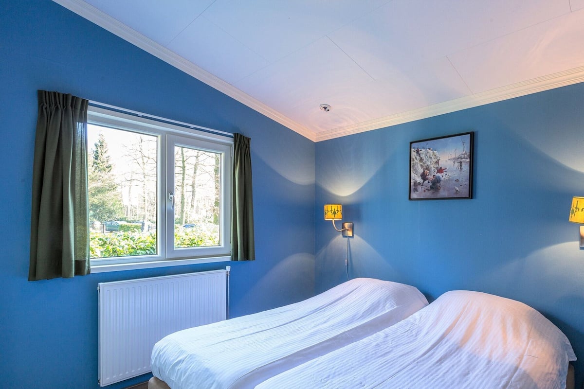 A room with two beds, a window with curtains, a radiator, and a wall-mounted lamp.