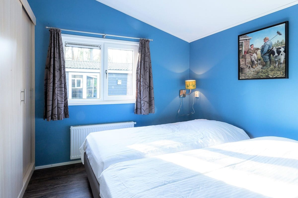 A bedroom with blue walls, a bed, a window with curtains, a radiator, and a wall-mounted lamp.
