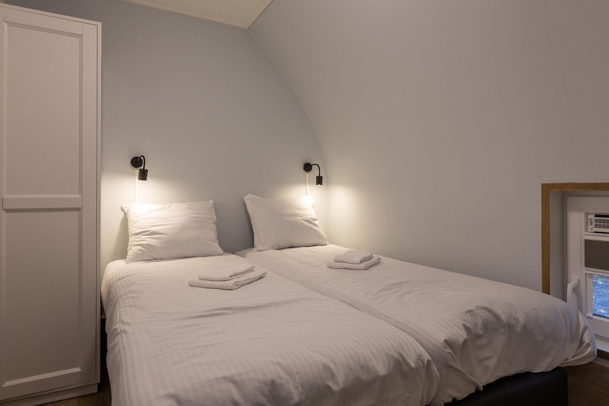 A bedroom with a sloped ceiling, two beds with white linens, two wall-mounted reading lights, and a window with an air conditioning unit.