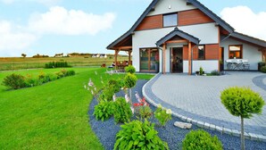 Una casa moderna de dos pisos con techo inclinado, un porche cubierto y un jardín ajardinado con diversas plantas y flores.