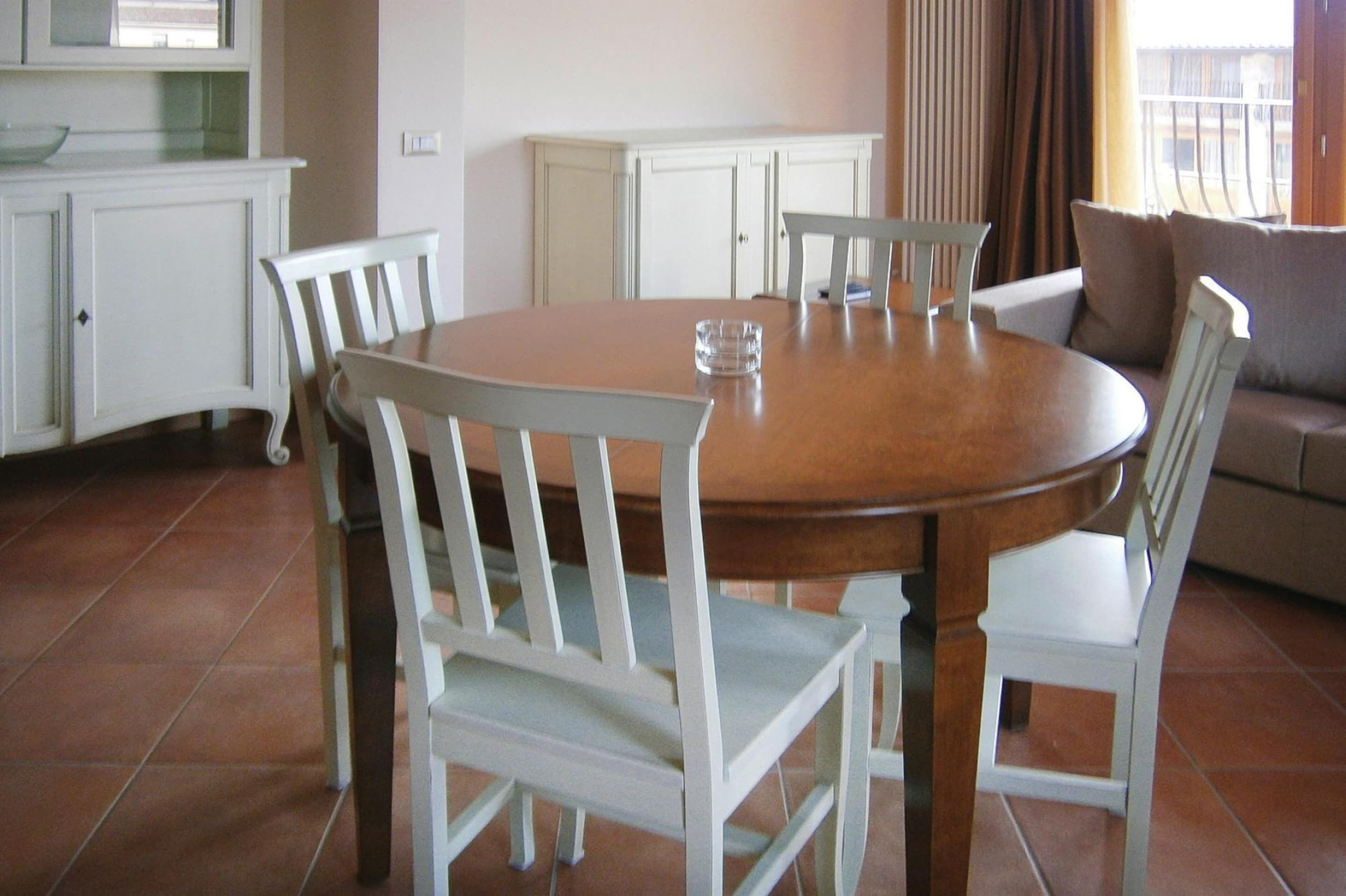 A dining area with a round wooden table surrounded by white chairs, a kitchen area with white cabinets, and a view of the outdoors through a window with curtains.