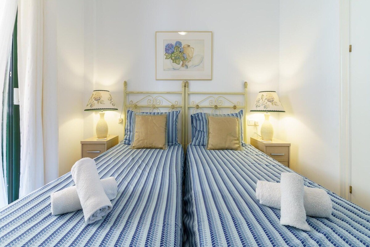A double bed with a striped blue and white bedspread, two bedside lamps, a framed picture above the bed, and rolled towels on the bed.