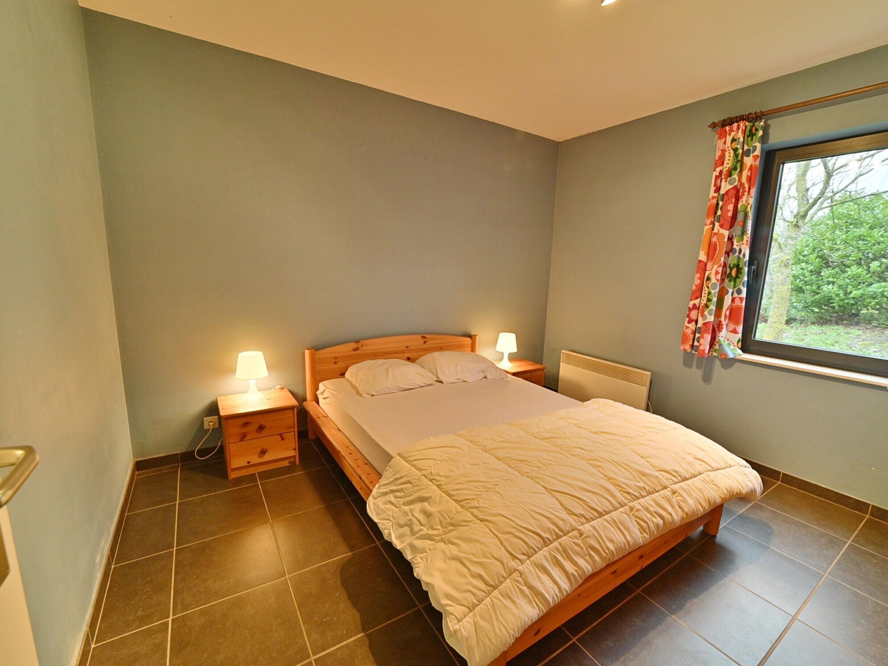 A bedroom with a bed, two bedside tables with lamps, a window with curtains, and a radiator.