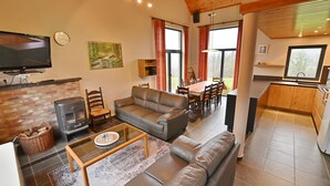 A living room with a fireplace, sofa, and television. There is a dining area with a table and chairs. The kitchen is visible with a window and a sink. - Modern Holiday Home in Somme-leuze With Garden (Somme-Leuze)