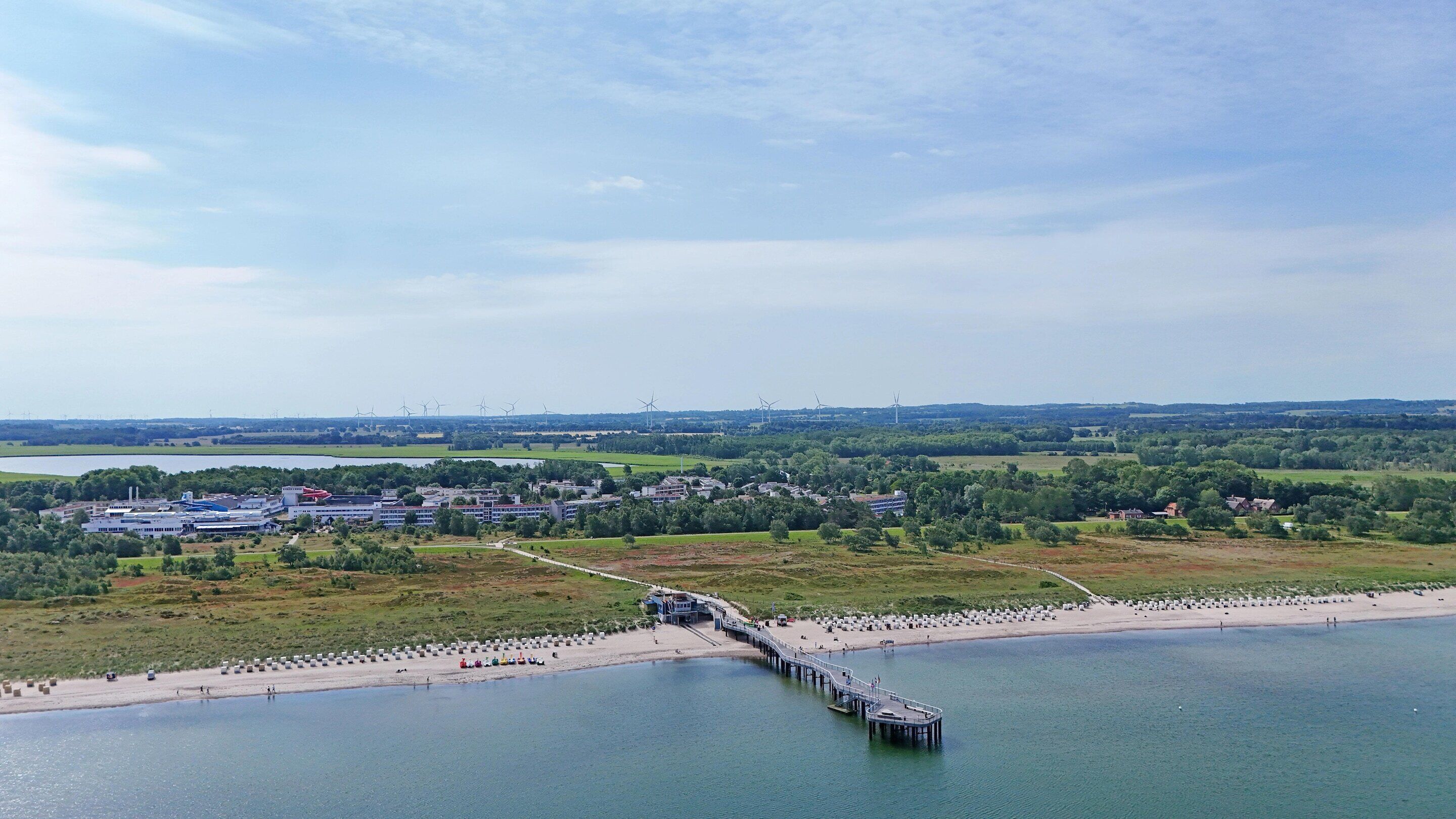 A coastal area with a pier extending into the water, a beach with sun loungers, and a backdrop of wind turbines and industrial buildings.