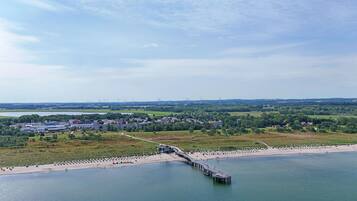 A coastal area with a pier extending into the water, a beach with sun loungers, and a backdrop of wind turbines and industrial buildings.