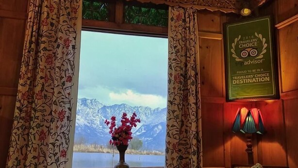 A room with a wooden ceiling, a chandelier, a window with a view of mountains, a framed award, and a floral-patterned sofa. - Kings Houseboats (Srinagar)