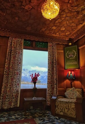 A room with a wooden ceiling, a chandelier, a window with a view of mountains, a framed award, and a floral-patterned sofa. - Kings Houseboats (Srinagar)