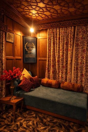 A cozy room with a wooden paneled wall, a framed certificate, a blue sofa, patterned curtains, and a floral vase. - Kings Houseboats (Srinagar)
