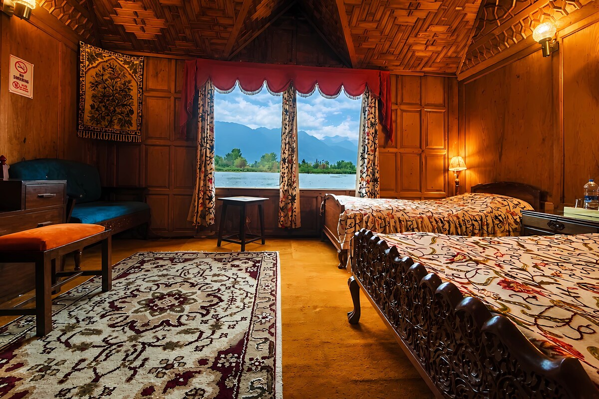 A room with a bed, a chair, a desk, a window with curtains, and a view of a lake and mountains.