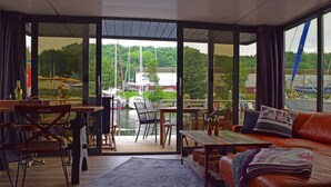 A modern living room with a large glass door leading to a deck, a leather sofa, a coffee table, and a dining area with chairs. - Houseboat Leni, Flensburg (Flensburg)