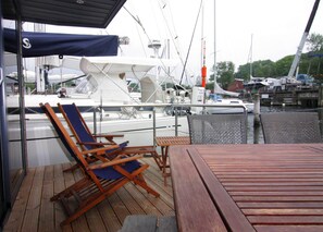 A covered deck with wooden lounge chairs and a table, overlooking a marina with boats. - Houseboat Leni, Flensburg (Flensburg)