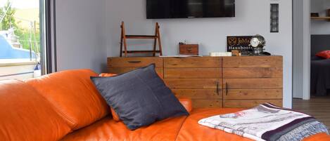 A living room with an orange leather sofa, a wooden dresser, and a flat-screen TV mounted on the wall.