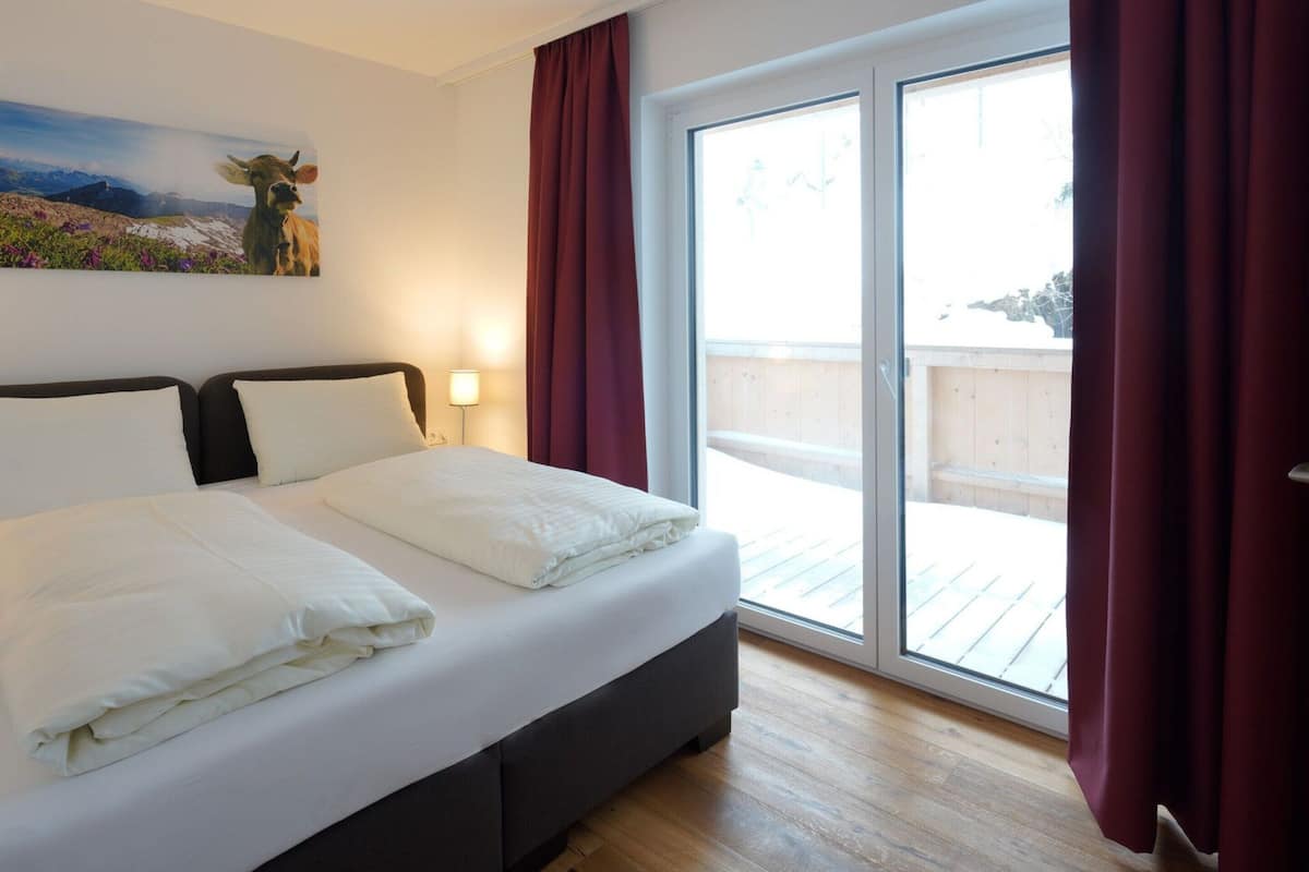 A hotel room with a bed, a painting on the wall, a window with curtains, and a view of the outside.