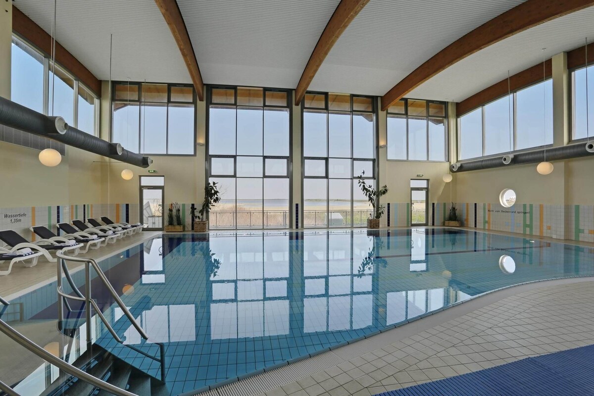 A large indoor swimming pool with clear windows, sun loungers, and a tiled floor.