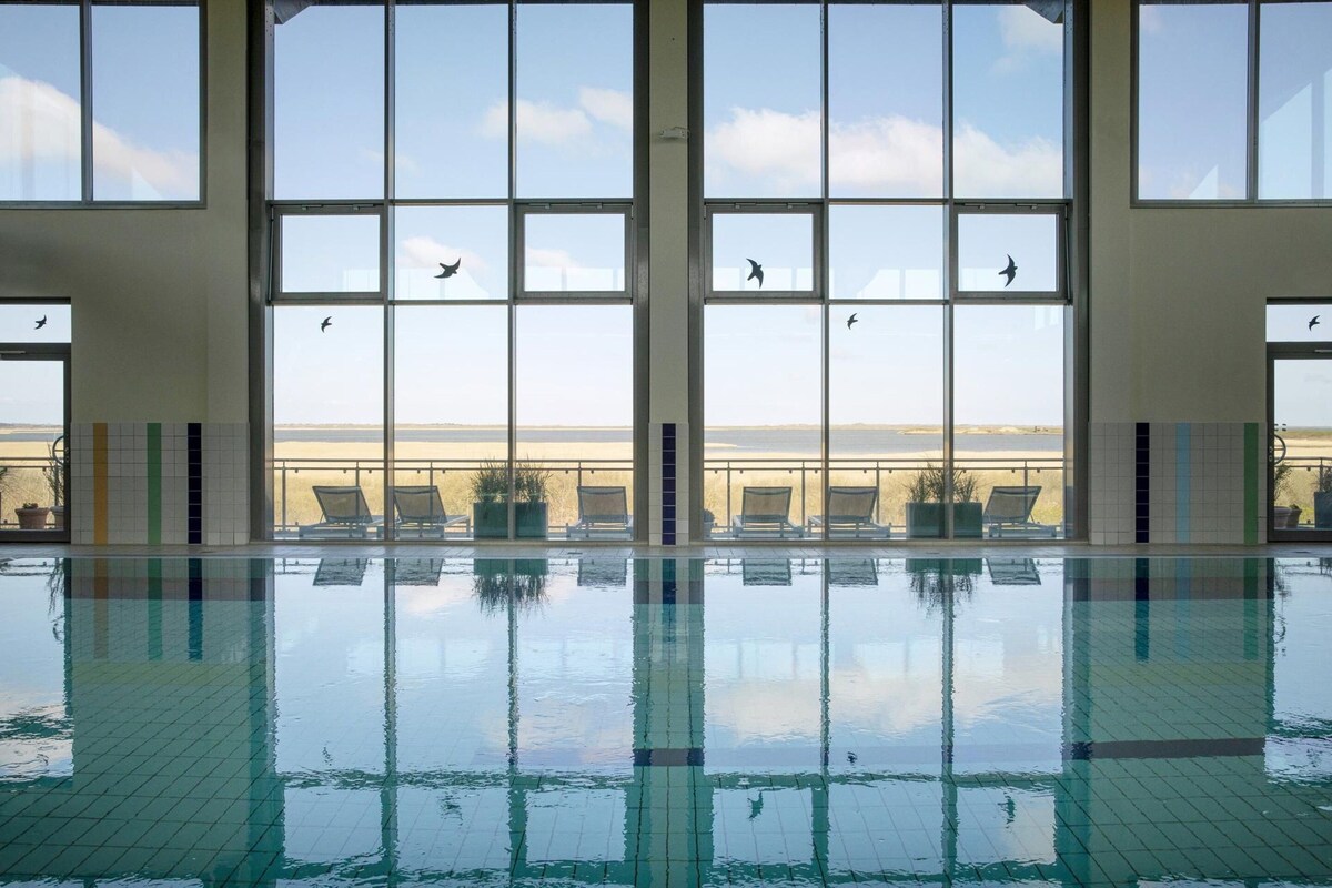 A large indoor swimming pool with clear glass walls, allowing views of the sky and birds outside.