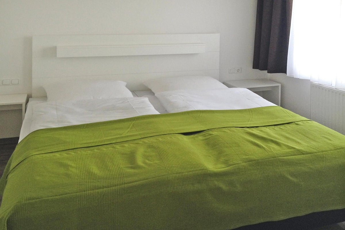 A hotel room with a bed, white headboard, a bedside table, and a window with curtains.