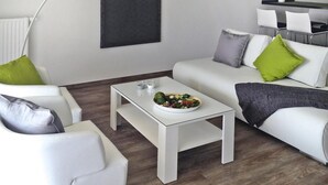 A modern living room with a white sofa, a coffee table with a bowl of fruit, and a dining area with a blackboard on the wall. - Apartments at Heidensee, Schwerin (Schwerin)