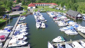 A marina with numerous boats docked in a calm water setting, surrounded by greenery and a large building with a red roof. - Apartments at Heidensee, Schwerin (Schwerin)