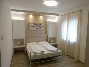 Um quarto com cama, mesas de cabeceira, um criado-mudo, uma obra de arte na parede e uma janela com cortinas.