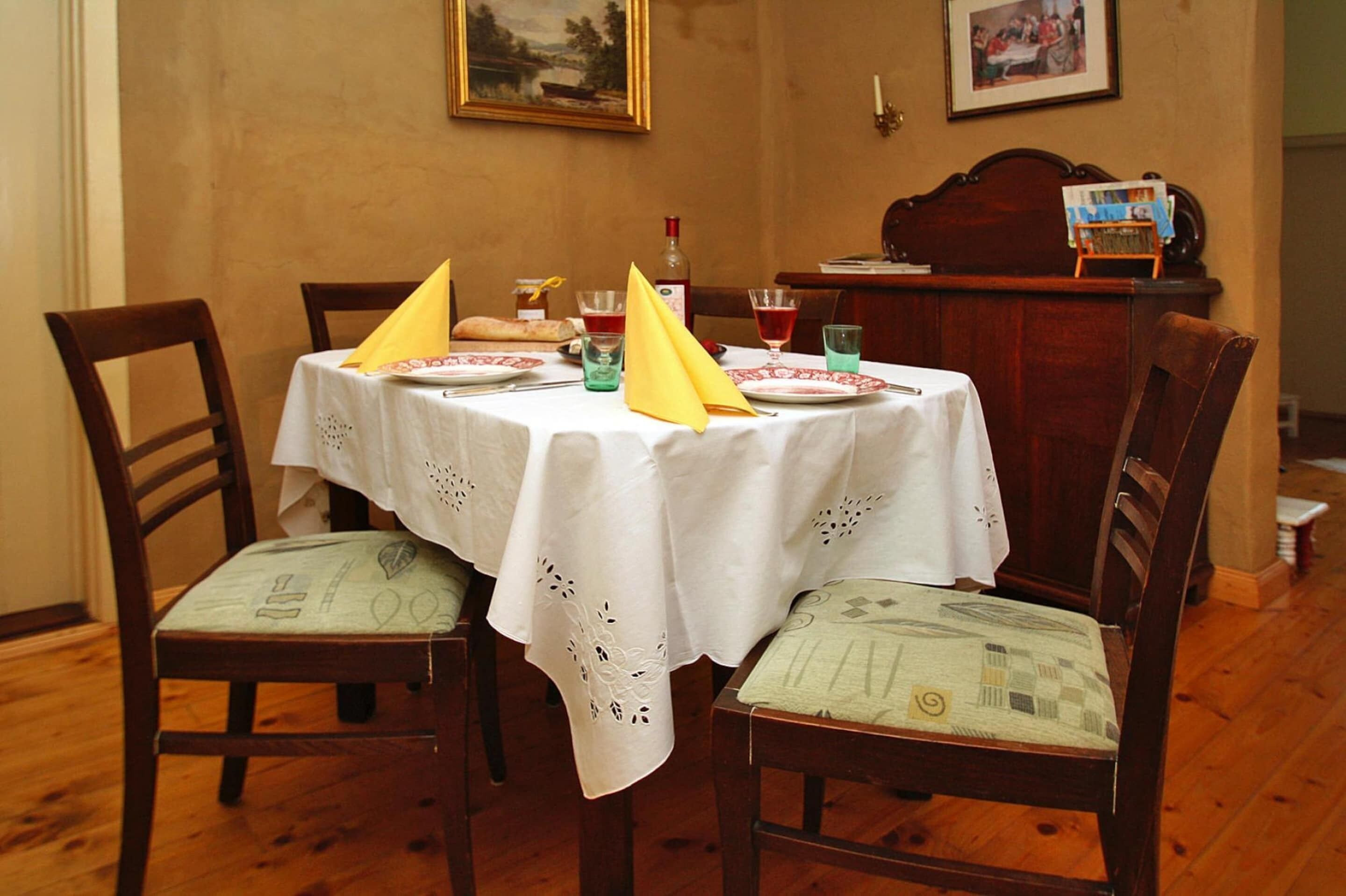 Une table à manger dressée avec une nappe blanche, quatre chaises et divers éléments de vaisselle, notamment des assiettes, des verres et une bouteille.
